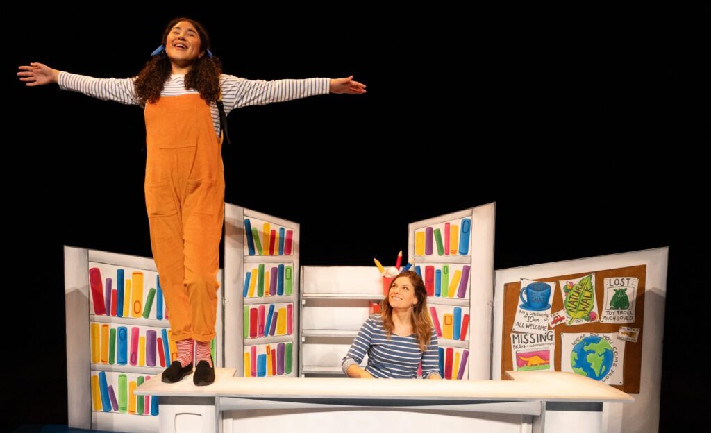 A picture of Luna Love Library Day production featuring Luna stood tall on a desk with arms out and singing and her mother sitting at the desk playing the piano.